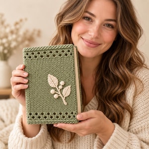 May include: A rectangular book cover made of sage green knitted yarn featuring a decorative openwork border at the top and bottom edges. A cream-coloured crocheted applique of a leafy branch with small round berries is centred on the front.