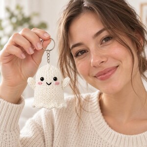 May include: A small, off-white knitted ghost keyring featuring two black bead eyes, a stitched black V-shaped mouth, and two small pink embroidered circles on the cheeks. The ghost is attached to a silver-toned metal split ring and chain.