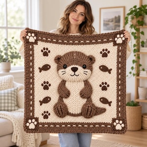 May include: A square, chunky knit blanket featuring a central 3D-style applique of a brown and cream otter. The cream-colored background is decorated with small brown paw prints and fish motifs, framed by a thick, dark brown border with paw prints.