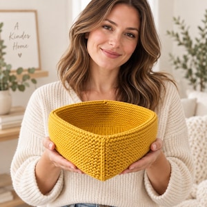 May include: A mustard yellow, heart-shaped basket crafted from thick, textured yarn. The basket features a dense, uniform knit pattern with a prominent vertical seam running down the centre of the front, creating a distinct, symmetrical shape.