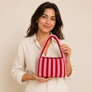 May include: A rectangular crochet handbag featuring vertical stripes in alternating shades of light pink, dark red, and bright red. The bag has a single, sturdy, arched handle constructed with the same striped pattern and a textured yarn finish.
