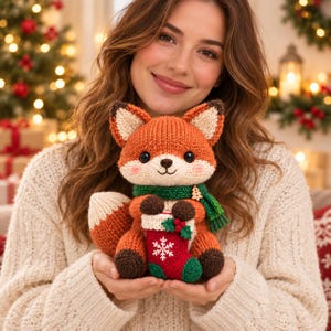 May include: A knitted fox plush toy featuring orange and white fur, dark brown paws, and a green scarf with a small wooden tree charm. The fox holds a miniature red and white Christmas stocking decorated with a white snowflake and green holly.
