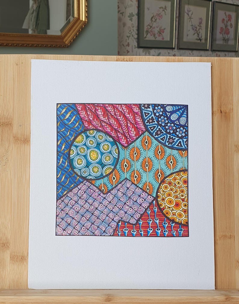 Peut inclure: Un dessin carr&eacute; sur papier blanc pr&eacute;sentant un motif g&eacute;om&eacute;trique vibrant compos&eacute; de plusieurs sections. Le design inclut des motifs complexes dessin&eacute;s &agrave; la main dans des tons de bleu, rouge, orange et jaune, avec des cercles et des lignes.