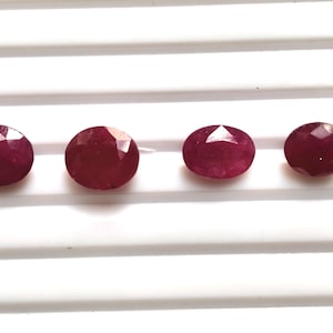 Puede incluir: Cuatro piedras preciosas rojas de forma ovalada y talladas en facetas, dispuestas en una fila horizontal. Cada piedra presenta una superficie pulida con múltiples facetas triangulares que reflejan la luz, mostrando un color carmesí profundo.