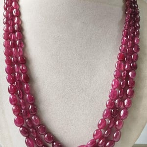 May include: A three-strand necklace composed of polished, oval-shaped, deep magenta-colored beads. The beads are strung in a multi-layered arrangement, creating a cascading effect around the neck. Each bead features a smooth, reflective surface.
