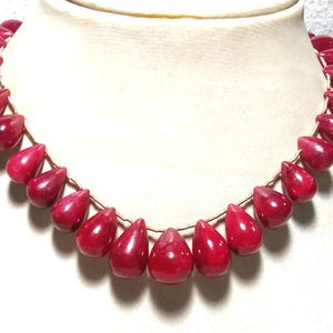Op de afbeelding: Een ketting met een reeks gepolijste, dieprode, traanvormige kralen geregen aan een dun, lichtgekleurd koord. De kralen zijn in een symmetrisch patroon gerangschikt, met de grootste traanvormige kraal in het midden geplaatst.