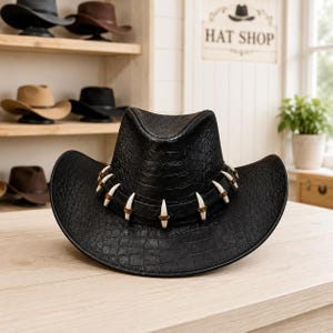 May include: A black cowboy hat with a textured crocodile skin pattern. The hat features a decorative band adorned with a series of white, curved, tooth-like accents held in place by small brown rings encircling the crown of the hat.
