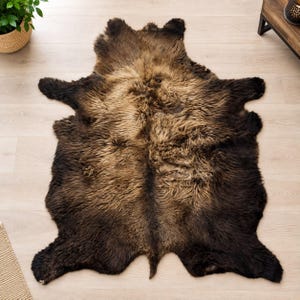 May include: A large, irregular-shaped animal hide rug featuring long, thick fur in a gradient of dark brown at the edges transitioning to a lighter, golden-brown tone toward the center, with a natural, organic silhouette and textured pile.