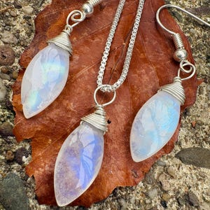 Op de afbeelding: Een sieradenset bestaande uit een ketting en twee bijpassende oorbellen, elk met een marquise-geslepen maansteen hanger gewikkeld in zilverdraad. De ketting bevat een zilveren boxketting, terwijl de oorbellen zijn voorzien van zilveren haken.