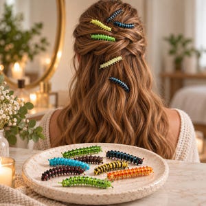 Op de afbeelding: Een verzameling haarclips in de vorm van rupsen in diverse kleuren, waaronder neongroen, geel, blauw, zwart en oranje. Verschillende clips liggen op een gespikkeld keramieken bord, terwijl andere in lang, golvend bruin haar zijn bevestigd.