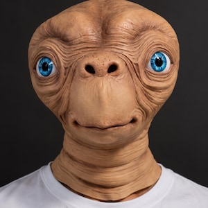 Pode incluir: Uma máscara de alienígena de cor bronzeada com uma cabeça grande, lisa e alongada, rugas proeminentes e olhos azul-brilhantes. A máscara apresenta um nariz largo e plano e uma boca pequena com lábios finos, sobre uma t-shirt branca lisa.