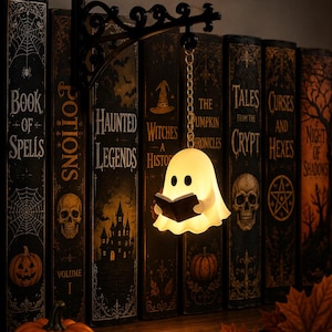 May include: A small, glowing white ghost figurine holding a black book, suspended from a black metal wall bracket by a gold-colored chain. The ghost has two black eyes and a smooth, rounded shape, emitting a soft light against a dark background.