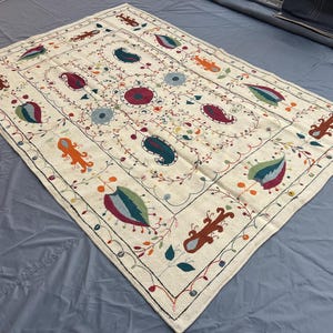 May include: A rectangular off-white textile featuring intricate hand-stitched embroidery. The design includes colorful paisley motifs, floral patterns, and stylized leaf shapes in shades of red, blue, green, and orange, all connected by thin, dark lines.