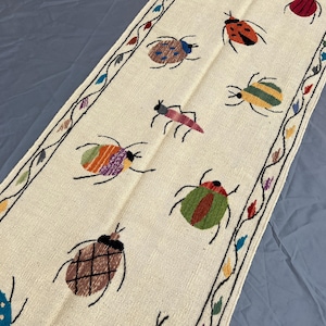 May include: A long, off-white table runner featuring embroidered, colorful beetles in various patterns and shades, including red, orange, green, blue, and brown. The runner is bordered by a thin, dark, winding vine pattern with small leaves.
