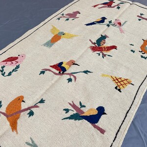 May include: A rectangular off-white fabric runner featuring embroidered birds in various colors including red, blue, yellow, orange, and pink. The birds are depicted perched on branches or in flight, with a thin black stitched border around the edge.