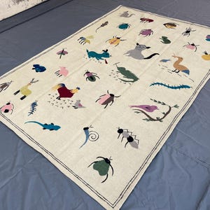 May include: A rectangular off-white textile rug featuring a repeating pattern of embroidered colorful animals and insects, including a rooster, a cat, a bird on a branch, and various beetles, all enclosed within a thin black stitched border.