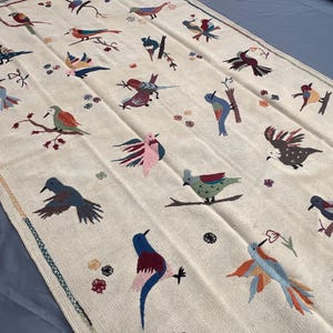 May include: A rectangular off-white textile featuring an embroidered pattern of various birds in flight and perched on branches. The birds are depicted in multiple colors including blue, red, pink, and brown, interspersed with small floral motifs.