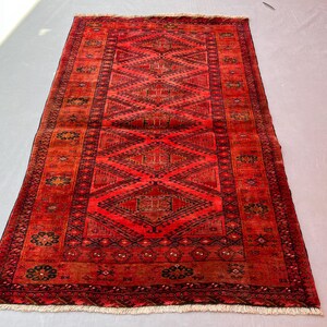 May include: A rectangular area rug featuring a vibrant red background with a repeating pattern of large black diamonds along the center. The border displays intricate geometric motifs and floral elements in black and dark red, finished with white fringe.