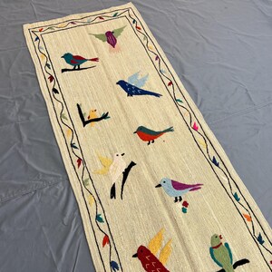 May include: A rectangular off-white table runner featuring colorful embroidered birds in various poses. The border consists of a thin black line with small embroidered leaves and flowers in red, blue, and green. The birds are depicted in shades of blue, red, yellow, and green.