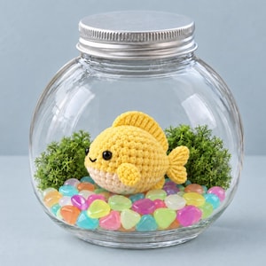 May include: A clear glass bowl with a silver lid containing a yellow crocheted fish toy, green moss clusters, and a layer of colorful, translucent pebbles at the bottom. The fish has black bead eyes and a small stitched smile.