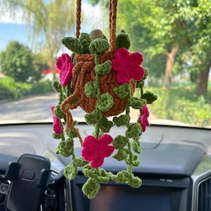 May include: A crocheted hanging plant ornament featuring a brown pot with green trailing vines and vibrant pink flowers. The item is suspended by a brown crocheted cord and includes a small wooden bead at the top of the pot for decoration.