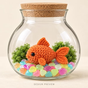 May include: A clear glass bowl with a cork lid containing a crocheted orange goldfish, green moss, and multicoloured translucent pebbles. The text DESIGN PREVIEW is printed at the bottom centre of the image.