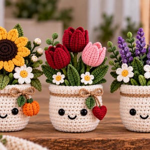 Puede incluir: Tres plantas en maceta tejidas a ganchillo con caras sonrientes. La primera tiene un girasol amarillo, la segunda tulipanes rojos y rosas, y la tercera lavanda morada. Cada maceta blanca tiene un lazo de cuerda y un pequeño adorno tejido.