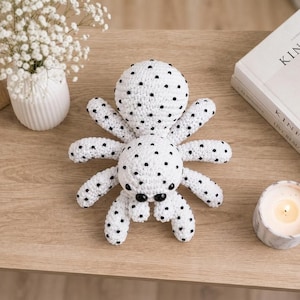 Puede incluir: Un peluche de araña tejido a ganchillo en color blanco con pequeños lunares negros en su cuerpo redondo y ocho patas. La araña tiene dos ojos negros grandes y prominentes situados en la parte frontal de la cabeza, con acabado de hilo.