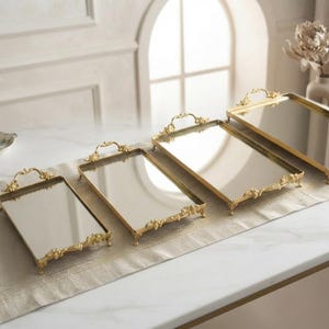Könnte beinhalten: Ein Set aus vier rechteckigen Spiegeltabletts mit goldfarbenen Metallrahmen. Jedes Tablett verfügt über verzierte Griffe und kleine Füße. Die Tabletts sind in einer Reihe angeordnet und nehmen von links nach rechts in ihrer Größe zu.