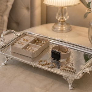 Può includere: Un vassoio rettangolare argentato a specchio con manici e piedini decorati. Contiene un organizer per gioielli in velluto beige con anelli e orecchini, uno smartwatch beige, due orecchini a cerchio dorati e una molletta per capelli con cristalli.