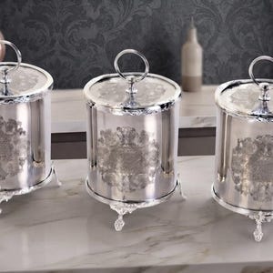 May include: Three cylindrical silver-toned canisters with lids, each featuring a circular handle on top and three ornate feet. The exterior of each canister is decorated with a detailed embossed pattern on the front, creating a reflective surface.