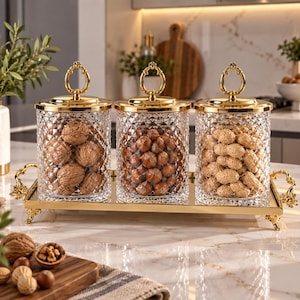 May include: Three clear glass canisters with diamond-patterned textures and gold-toned lids featuring oval handles. The canisters sit on a rectangular gold-toned mirrored tray with ornate handles and feet, filled with walnuts, hazelnuts, and peanuts.