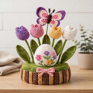 Op de afbeelding: Een gehaakt arrangement met een centraal wit ei versierd met kleine kleurrijke bloemen, omringd door vier tulpen in paars, roze, geel en wit. Een paarse en roze vlinder zit erboven, alles op een ronde groene basis met een roze strik.