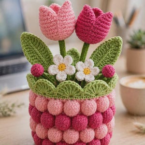 May include: A crochet flower arrangement featuring two tulips in light and dark pink, two small white flowers with yellow centers, and two pink spheres, all set in a textured, cylindrical basket made of alternating rows of pink bobble stitches.