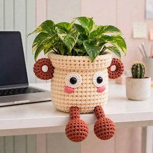 Op de afbeelding: Een crèmekleurige gehaakte plantenpotcover met een karakterontwerp met twee bruine ronde oren, twee grote witte ogen met zwarte pupillen, roze geborduurde wangen en twee bungelende bruine benen met ronde voeten die een plant vasthoudt.