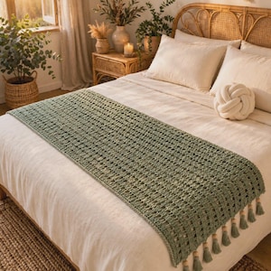 May include: A sage green rectangular crochet bed runner featuring an openwork lattice pattern and a decorative fringe edge with small wooden beads attached to each tassel, draped horizontally across a neutral-colored bedspread.