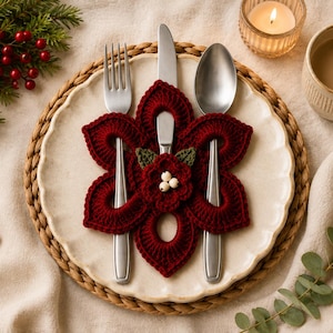 May include: A dark red crocheted cutlery holder shaped like a flower with six petals, featuring a small central floral accent with three white beads and two green leaves, holding a silver fork, knife, and spoon on a cream-coloured ceramic plate.
