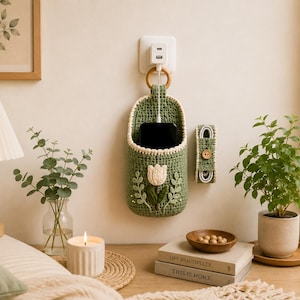 May include: A sage green crocheted wall-hanging phone holder featuring a cream-colored tulip appliqué and a wooden ring for hanging. Beside it is a matching small rectangular cord organizer strap secured with a single wooden button.