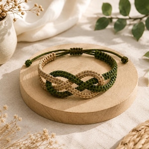 May include: A macrame bracelet featuring an infinity knot design crafted from intertwined beige and dark green cord. The bracelet includes an adjustable sliding knot closure with two thin green cords extending from the back, creating a secure fit.