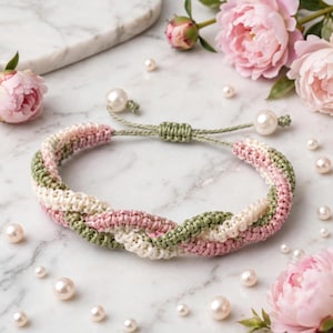 Puede incluir: Una pulsera de cordón trenzado con hebras entrelazadas en color verde salvia, rosa suave y crema. La pulsera incluye un cierre de nudo deslizante ajustable terminado con dos cuentas de perlas blancas en los extremos de los cordones verdes.