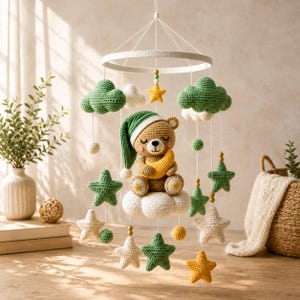 May include: A crochet baby mobile featuring a central brown bear wearing a green nightcap and holding a yellow crescent moon. The bear sits on a white cloud, surrounded by hanging green and white clouds, yellow and green stars, and small spheres.