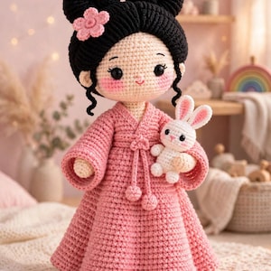 May include: A handmade crochet doll with black yarn hair styled in two buns, adorned with a small pink flower. The doll wears a long-sleeved pink dress with a tied waist and holds a small white crochet rabbit toy. It has rosy cheeks and black eyes.