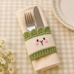 May include: A cream-coloured cloth napkin folded and held by a crocheted sleeve featuring a scalloped green top edge, a cream centre with an embroidered face, and a green bottom border, containing a stainless steel fork and a butter knife.