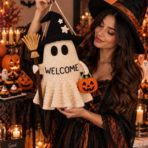 Op de afbeelding: Een gehaakte spookdecoratie met een zwarte heksenhoed met witte sterren, die een bruine bezem en een kleine oranje pompoen vasthoudt. Het gebroken witte spook heeft zwarte ovale ogen en het woord WELCOME geborduurd in zwarte draad.