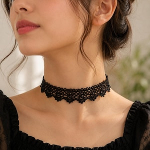 Puede incluir: Un collar tipo gargantilla de encaje negro con un intrincado patrón tejido y pequeños bordes festoneados en la parte inferior. El diseño consiste en un delicado trabajo de hilo que forma una estructura de red geométrica.