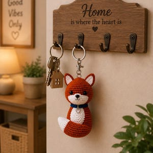 May include: A wooden wall-mounted key holder with four metal hooks, featuring the engraved text "Home is where the heart is" and a heart icon. A crochet fox keychain with a blue collar and a small heart charm hangs from one of the hooks.