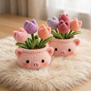 May include: Two crocheted pig-shaped pots containing colorful yarn tulips. Each pot is light pink with small ears, black bead eyes, and a snout. The tulips are in shades of pastel pink, purple, and orange, with green crocheted stems and leaves.