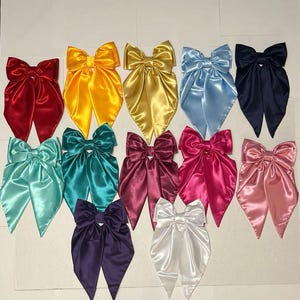 Puede incluir: Una colección de doce lazos para el cabello de satén en varios colores, incluyendo rojo, amarillo, dorado, azul claro, azul marino, verde azulado, burdeos, magenta, rosa, morado y blanco. Cada lazo tiene un acabado liso y brillante.