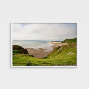 Könnte beinhalten: Ein gerahmtes Landschaftsfoto zeigt einen breiten Sandstrand, der von grünen, grasbewachsenen Hügeln unter einem bewölkten Himmel gesäumt wird. Die Ansicht zeigt die Küste am Ozean, wobei der nasse Sand das Licht reflektiert und die Hügel abfallen.