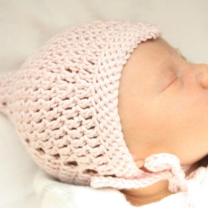 Könnte beinhalten: Eine rosa gehäkelte Babyhaube mit einem gekräuselten Rand. Die Haube wird von einem schlafenden Baby getragen.